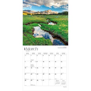 May include: A 2027 wall calendar for the month of March featuring a photograph of a green meadow with a winding stream reflecting the sky and distant snow-capped mountains. The calendar grid displays dates from the 1st to the 31st of March.