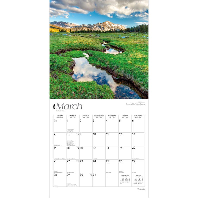 May include: A 2027 wall calendar for the month of March featuring a photograph of a green meadow with a winding stream reflecting the sky and distant snow-capped mountains. The calendar grid displays dates from the 1st to the 31st of March.