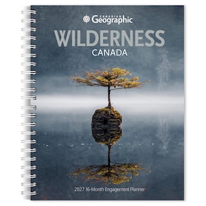 May include: A spiral-bound 2027 16-Month Engagement Planner titled Canadian Geographic WILDERNESS CANADA. The cover features a photograph of a small tree growing on a rock in the middle of a calm, misty lake, with the tree reflected in the water.