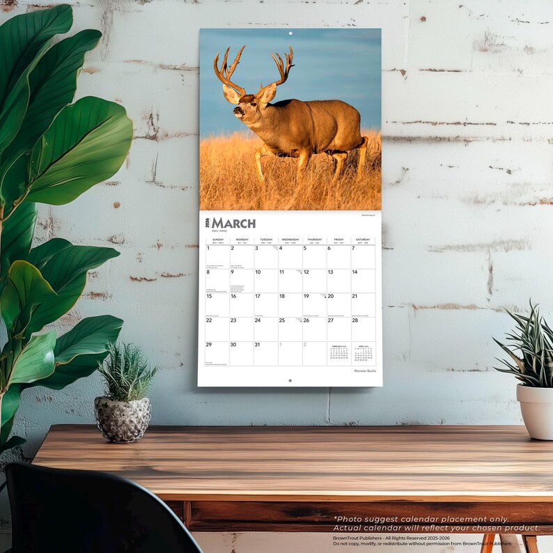 May include: A wall calendar featuring a majestic deer with large antlers in a field of tall grass under a blue sky. The calendar is open to March, with a white grid of dates. The calendar is hanging on a white brick wall.