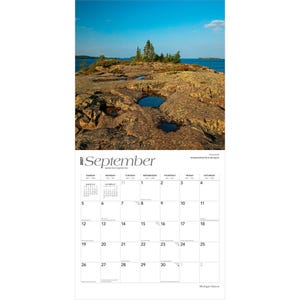 May include: A 2027 wall calendar for the month of September featuring a photograph of a rocky shoreline with small pools of water and evergreen trees against a blue sky. The calendar grid displays dates from Sunday through Saturday with black text.