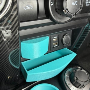May include: A close-up of a car's interior with two turquoise plastic storage compartments. The compartments are located in the center console, below the audio and HDMI ports. The compartments are designed to hold small items, such as phones, wallets, or keys.