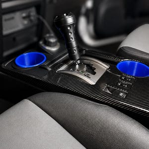 May include: A black carbon fiber automotive center console featuring a gear shift lever with a cylindrical black knob, two bright blue circular cup holder inserts, and integrated switches for vehicle controls positioned near the gear shifter.