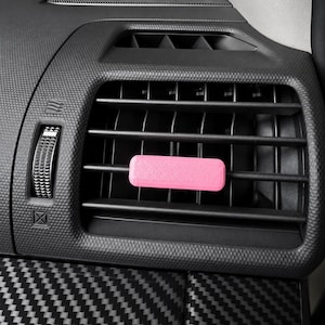 Könnte beinhalten: Ein rechteckiger, rosafarbener Lufterfrischer-Clip, der an den horizontalen Lamellen einer schwarzen Kunststoff-Lüftungsdüse im Auto befestigt ist. Die Lüftung hat eine strukturierte Oberfläche, ein gerändeltes Einstellrad und ein Carbonmuster.