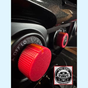 May include: Close-up of a car's dashboard featuring red control knobs. The knobs are textured and have white lettering indicating their functions. A logo for "5th Gen Runner" is visible in the lower right corner. The dashboard is black and glossy.