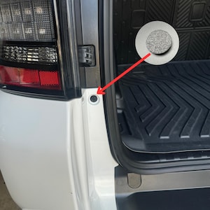 Puede incluir: Un coche blanco con un parachoques trasero negro y una alfombra de carga de plástico negro. Un tornillo plateado es visible en el parachoques trasero.