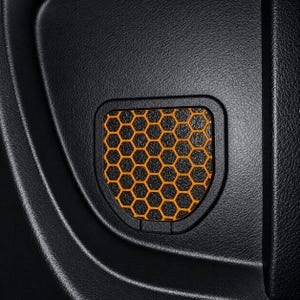 May include: A black automotive interior panel featuring a recessed, shield-shaped insert. The insert displays a vibrant orange honeycomb pattern set against a textured black background, framed by a smooth, rounded black border.
