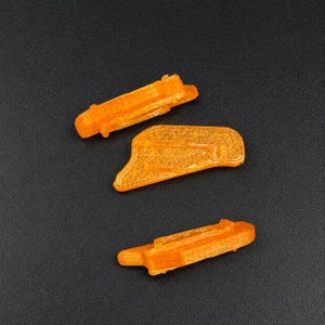 May include: Three translucent orange plastic components with textured surfaces. Two pieces are elongated with small protruding tabs, while the third piece is wider and features a distinct angular shape with a raised border along its edge.