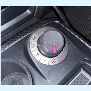 Może przedstawiać: Zbliżenie wnętrza pojazdu z okrągłym pokrętłem sterowania z ustawieniami L4, H4 i H2. Pokrętło otoczone jest srebrnym pierścieniem i czarną, teksturowaną powierzchnią. Na pokrętle znajduje się różowa naklejka.