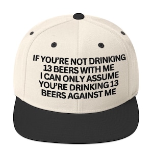 May include: A cream-colored baseball cap with a black brim and the text "IF YOU'RE NOT DRINKING 13 BEERS WITH ME I CAN ONLY ASSUME YOU'RE DRINKING 13 BEERS AGAINST ME" in black. The cap has a flat brim and a button on top.