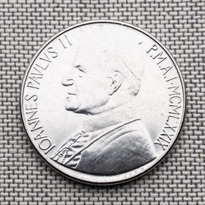 Puede incluir: Una moneda conmemorativa circular de color plateado que presenta un retrato de perfil del Papa Juan Pablo II mirando hacia la izquierda. La inscripción dice IOANNES PAVLVS II a la izquierda y P.M.A.I. MCMLXXIX en el borde derecho de la moneda.