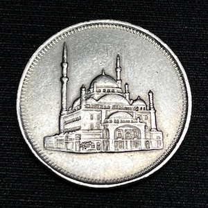 Puede incluir: Una moneda de plata con una representación detallada de un edificio con cúpulas y minaretes. El borde de la moneda tiene un patrón texturizado. La moneda está sobre un fondo negro.