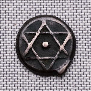 Puede incluir: Una moneda o ficha circular de metal oscuro con un diseño en relieve de una estrella de seis puntas y un pequeño punto centrado en su interior. La superficie metálica muestra una pátina envejecida con reflejos metálicos claros.