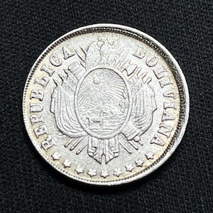 May include: A silver coin with intricate details, featuring the words "REPUBLICA BOLIVIANA" around the edge. The coin's center showcases an emblem with an eagle, flags, and a landscape scene. The coin is set against a dark background.