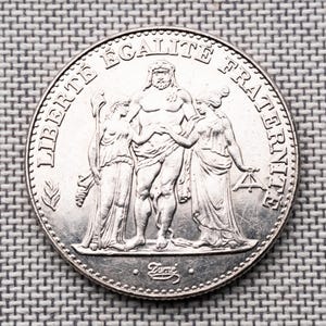 May include: A silver-coloured French coin featuring a central male figure flanked by two female figures, with the text LIBERTÉ ÉGALITÉ FRATERNITÉ arched across the top and the signature Dupré inscribed at the bottom edge of the circular metal piece.