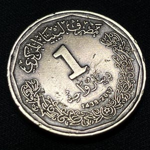 Puede incluir: Una moneda plateada con escritura árabe y el número 1 en el centro. La moneda presenta diseños florales ornamentados en los bordes y la fecha 1439-2017. El fondo es negro.