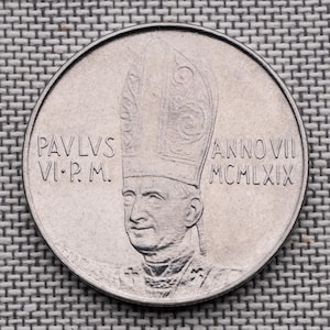 Może przedstawiać: Okrągła, srebrzysta moneta okolicznościowa z wypukłym portretem papieża Pawła VI w mitrze. Napis PAVLVS VI P. M. znajduje się po lewej stronie, a ANNO VII MCMLXIX po prawej stronie centralnej postaci na awersie monety.