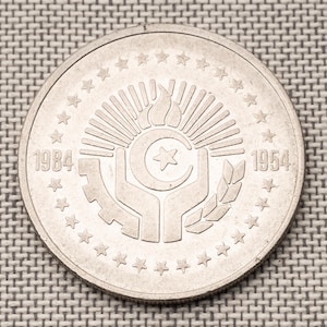 May include: A silver coin featuring a central emblem with a crescent moon, star, and flame, surrounded by a gear, hands, and wheat. The coin is ringed with stars and the dates 1984 and 1954 are visible.