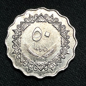 Puede incluir: Una moneda plateada con borde festoneado, con escritura árabe y un diseño central de un número y espigas de trigo. La moneda tiene un borde ornamentado sobre un fondo oscuro.