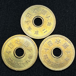 May include: Three Japanese 5-yen coins with a central hole and embossed Japanese characters. The coins are gold-colored and feature a design with text and symbols. The coins are arranged on a black surface.