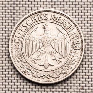 Puede incluir: Una moneda de plata de 1928 con el texto "DEUTSCHES REICH" y un emblema de águila. La moneda tiene un diseño detallado con una corona alrededor del águila y texto. La moneda es circular.