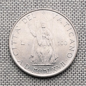 Puede incluir: Una moneda de 100 liras de la Ciudad del Vaticano de color plateado con un relieve central de una figura sentada con la mano levantada. El texto dice CITTA DEL VATICANO, L. 100, y la fecha MCMLXVII, con las iniciales G.P.F. visibles abajo.