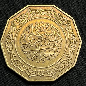 Puede incluir: Moneda octogonal dorada con escritura árabe ornamentada en el centro. La moneda tiene un borde decorativo con diseños arremolinados. La moneda está sobre un fondo negro.