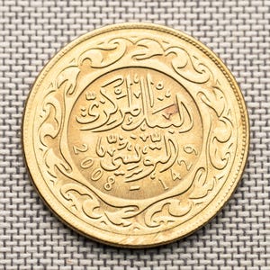 Puede incluir: Una moneda dorada con escritura árabe y los números 2008 y 1429. La moneda tiene un diseño de borde ornamental y arremolinado. La moneda está sobre una superficie texturizada de color gris claro.