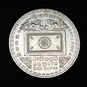 Puede incluir: Una moneda de plata de Sri Lanka con grabados detallados. Muestra dos leones flanqueando un emblema rectangular, con las inscripciones "SRI LANKA" y "ONE RUPEE". La moneda tiene un borde con diseños.