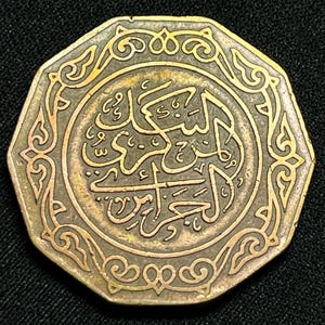 Puede incluir: Moneda de color bronce con escritura árabe en el centro y diseños ornamentales, similares a flores, alrededor del borde. La moneda tiene forma octogonal y está sobre un fondo oscuro.