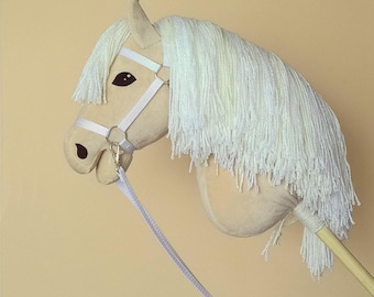 May include: A plush toy horse head in cream and white. The horse has a long, flowing white mane and a cream-colored head with black eyes and a white bridle. A wooden stick extends from the bottom, and a white leash is attached.