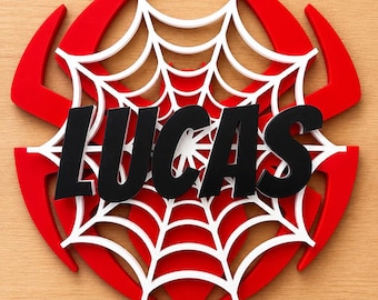 May include: A red, white, and black wall decoration featuring the name "LUCAS" in bold black letters. The design includes a red outer shape with white spiderweb detailing, creating a superhero-themed aesthetic.