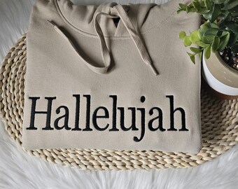 Può includere: Una felpa con cappuccio beige con un cordoncino nero e la parola "Hallelujah" ricamata in nero sul davanti.