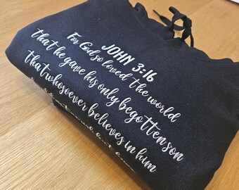 Può includere: Una felpa con cappuccio blu navy piegata con scritta bianca. Il testo recita "JOHN 3:16" seguito da un versetto biblico. La felpa con cappuccio ha un cordino ed è realizzata in un materiale morbido.