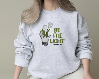 Op de afbeelding: Een lichtgrijs sweatshirt met een afbeelding van een gloeilamp met witte bloemen en de tekst "BE THE LIGHT" in het groen. De tekst "Matthew 5:14" staat eronder. Het sweatshirt heeft lange mouwen.