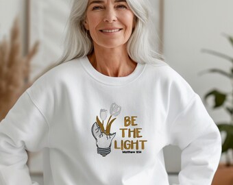 Op de afbeelding: Wit sweatshirt met een afbeelding van een kapotte gloeilamp met bloemen en de tekst "BE THE LIGHT Matthew 5:14" in goud. Het sweatshirt heeft een ronde hals.