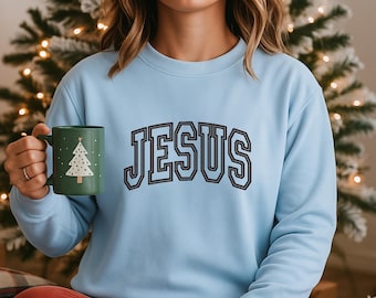 Può includere: Una felpa azzurra con la scritta "JESUS" in un font in stile collegiale. La felpa è abbinata a pantaloni rossi. Una tazza verde con un disegno di albero di Natale bianco è tenuta in mano.