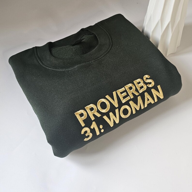 K&ouml;nnte beinhalten: Ein dunkelgr&uuml;ner Pullover mit goldener Stickerei, auf dem "Spr&uuml;che 31: Frau" steht.