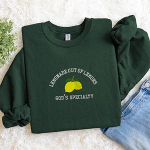 May include: A dark green sweatshirt with the embroidered text "LEMONADE OUT OF LEMONS" above a lemon graphic and the text "GOD'S SPECIALTY" below. The sweatshirt is made of a soft material.