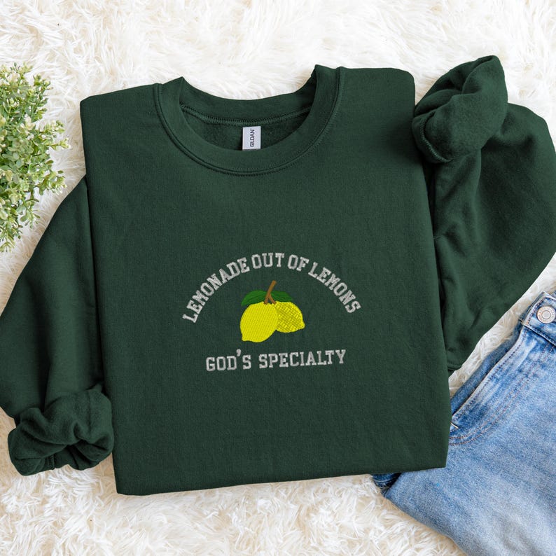 May include: A dark green sweatshirt with the embroidered text "LEMONADE OUT OF LEMONS" above a lemon graphic and the text "GOD'S SPECIALTY" below. The sweatshirt is made of a soft material.