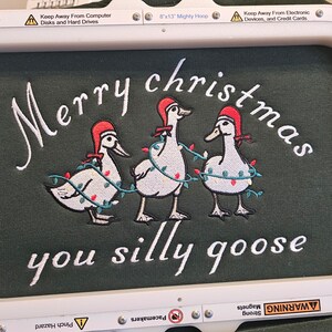 May include: Embroidered design on a green fabric with three white geese wearing red Santa hats and Christmas lights. The text "Merry christmas you silly goose" is embroidered below the geese.