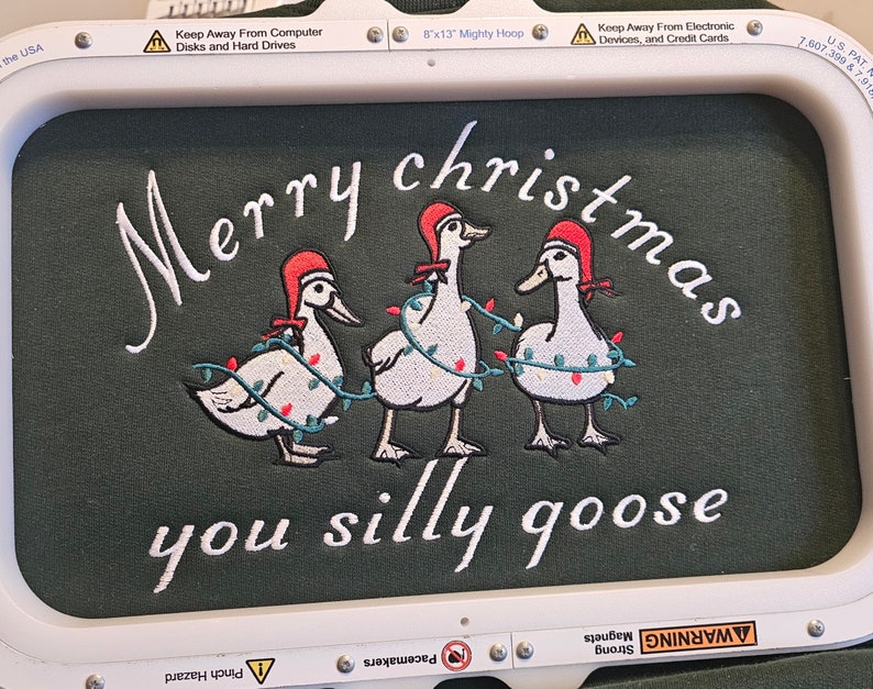 May include: Embroidered design on a green fabric with three white geese wearing red Santa hats and Christmas lights. The text "Merry christmas you silly goose" is embroidered below the geese.