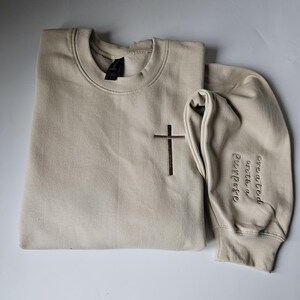 K&ouml;nnte beinhalten: Ein beiger Pullover mit einem braunen gestickten Kreuz auf der Vorderseite und den Worten "geschaffen mit einem Zweck" auf dem &Auml;rmel.