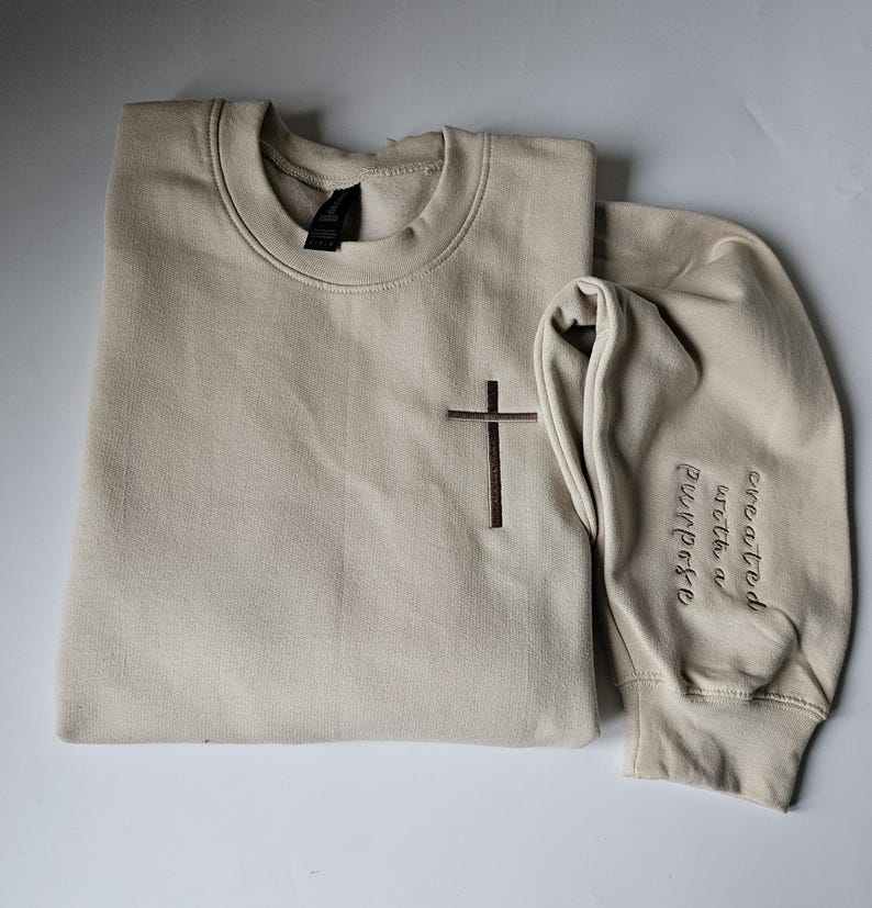 K&ouml;nnte beinhalten: Ein beiger Pullover mit einem braunen gestickten Kreuz auf der Vorderseite und den Worten "geschaffen mit einem Zweck" auf dem &Auml;rmel.