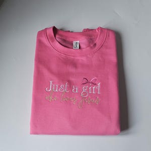 Op de afbeelding: Een gevouwen roze sweatshirt met de geborduurde tekst "Just a girl who loves Jesus" in witte en gouden draad. Een kleine strik is geborduurd boven het woord "girl".