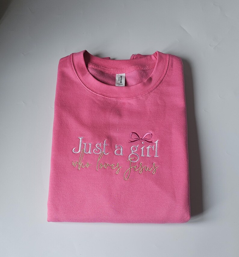 Op de afbeelding: Een gevouwen roze sweatshirt met de geborduurde tekst "Just a girl who loves Jesus" in witte en gouden draad. Een kleine strik is geborduurd boven het woord "girl".