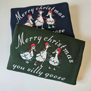 May include: Two sweatshirts with a Christmas theme. The top sweatshirt is navy blue with white text that says "Merry Christmas" and a line of geese wearing Christmas lights. The bottom sweatshirt is dark green with white text that says "Merry Christmas you silly goose" and a line of geese wearing Christmas lights.