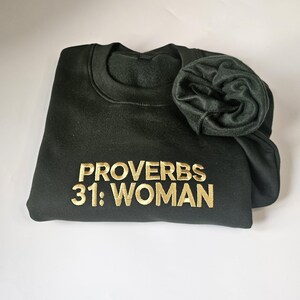 K&ouml;nnte beinhalten: Ein dunkelgr&uuml;ner Pullover mit dem Text "Spr&uuml;che 31: Frau" in goldener Stickerei.