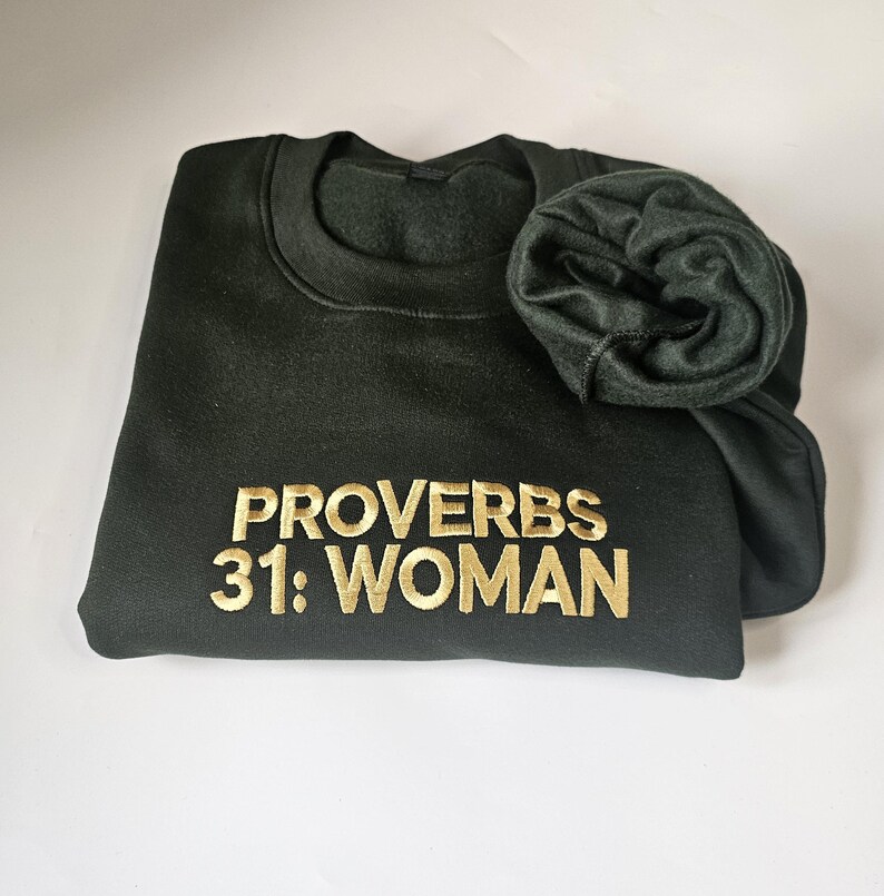 K&ouml;nnte beinhalten: Ein dunkelgr&uuml;ner Pullover mit dem Text "Spr&uuml;che 31: Frau" in goldener Stickerei.
