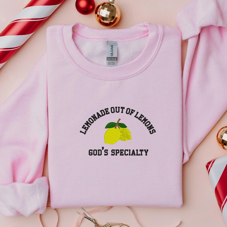 May include: A light pink sweatshirt with the embroidered text "LEMONADE OUT OF LEMONS GOD'S SPECIALTY" above a lemon graphic. The sweatshirt is surrounded by holiday decorations, including ornaments and a candy cane.